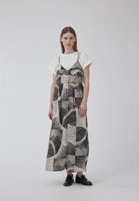 Robe maxi noire et crème avec des motifs abstraits, fines bretelles et boutonnage sur le devant, associée à un t-shirt blanc à manches courtes et des chaussures noires.