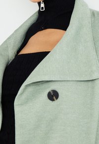 Chaqueta abotonada verde claro con una tela texturizada, un gran cuello y un cuello de tortuga negro acanalado debajo que cuenta con una cremallera plateada.