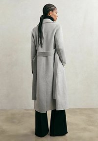 Siv volnen plašč z pasom v pasu, krojenim silhuetom in dolgimi rokavi; kombiniran s širokimi črnimi hlačami. Gladek tekstura, minimalističen dizajn.