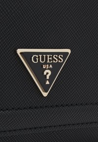 Černý texturovaný materiál s trojúhelníkovým logem v zlaté a černé barvě s nápisem „GUESS USA“ a otazníkem. Jednoduchý a geometrický design.
