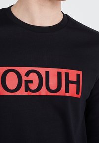Czarny t-shirt z okrągłym dekoltem, z wyraźnym czerwonym prostokątnym nadrukiem z napisem "HUGO" czarnymi literami. Gładka faktura.