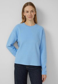 Frau mit hellbraunem Haar, die einen hellblauen Langarmpullover und dunkle Jeans trägt, steht vor einem schlichten grauen Hintergrund.