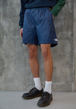 Marineblaue, leichte Shorts mit elastischem Bund und seitlichen Taschen; mit einem weißen Logo am linken Bein. Kombiniert mit schwarzen Schuhen und weißen Socken.