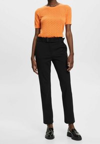 Orange stickad kortärmad topp med ett texturerat mönster, matchad med svarta skräddarsydda byxor och glansiga svarta loafers.