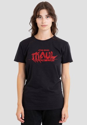 Junge Frau mit langen braunen Haaren, die ein schwarzes Star Wars Maul Shadow Lord T-Shirt trägt, steht vor einem schlichten grauen Hintergrund.
