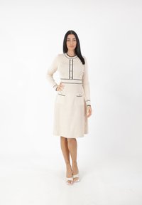 Beiges Kleid mit langen Ärmeln, Rundhalsauschnitt, strukturierter Stoff, schwarzen Akzenten, Knöpfen in der Mitte und zwei vorderen Taschen.