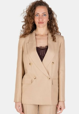Donna con capelli ricci che indossa un blazer doppiopetto beige sopra una maglia nera in pizzo, in piedi su sfondo bianco uniforme.