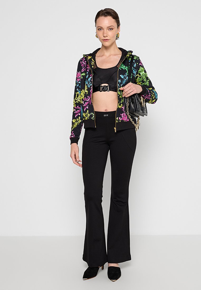 Wielokolorowa kwiatowa hoodie na zamek, czarny crop top z klamrą, czarne spodnie dzwony, czarna torebka i szpilki. Gładkie tekstury w całym zestawie.