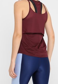 Top da allenamento color bordeaux con texture a coste, design a strati e spalline nere; abbinato a leggings blu e navy.