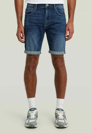 Homme portant un short en jean bleu retroussé, des chaussettes blanches et des chaussures de course grises, debout devant un fond uni.