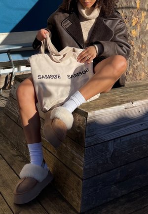 Femme assise en plein air sur un banc en bois, tenant un sac cabas beige avec le texte « SAMSØE SAMSØE », portant une veste marron, des chaussettes blanches et des chaussons doublés de fourrure.