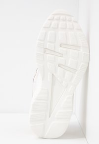 Semelle d'une chaussure de sport blanche montrant une bande de roulement texturée avec des motifs rectangulaires et triangulaires sur un fond blanc uni.