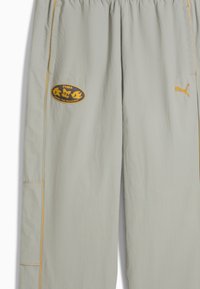 Lys grå bukser lavet af tekstureret stof, der har et gult logo-lap og gule side-striber. Inkluderer lomme detalje på venstre side.