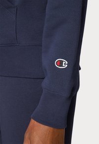 Marineblå sweatshirtstoff med ribbestrikkede mansjetter, med et hvitt og rødt broderi av logoen på venstre erme. Glatt tekstur, avslappet passform.