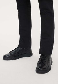 Black leather sneakers with textured side panels, chunky rubber soles, and traditional lacing. Paired with tapered black trousers.
