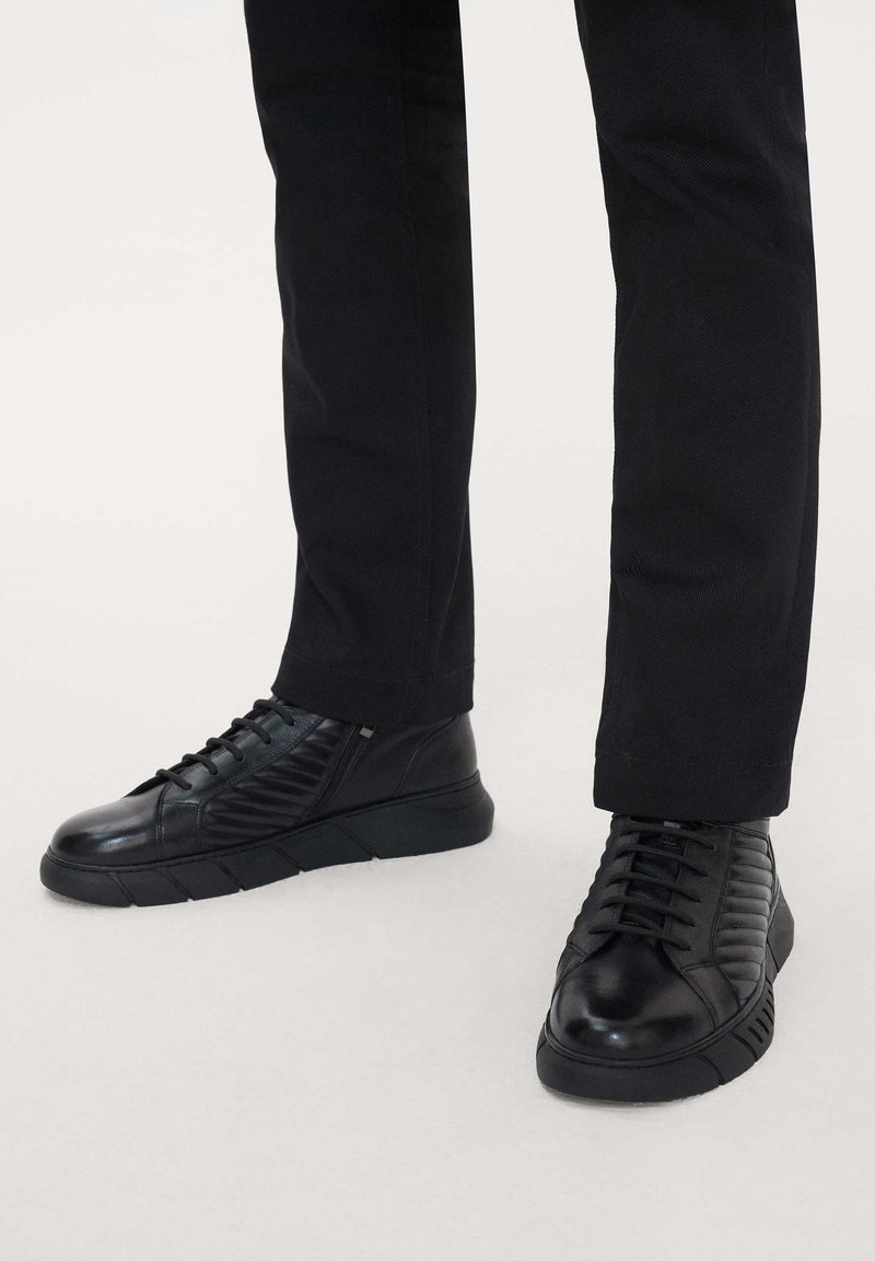 Black leather sneakers with textured side panels, chunky rubber soles, and traditional lacing. Paired with tapered black trousers.