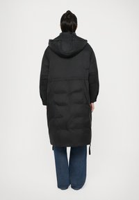 Doudoune noire avec capuche, design long, taille cintrée et détails de couture visibles. Portée sur un jean large.