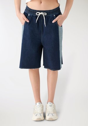 Persona che indossa pantaloncini di jeans bicolore fino al ginocchio con vita elastica e coulisse e sneakers bianche, in piedi con le mani nelle tasche.
