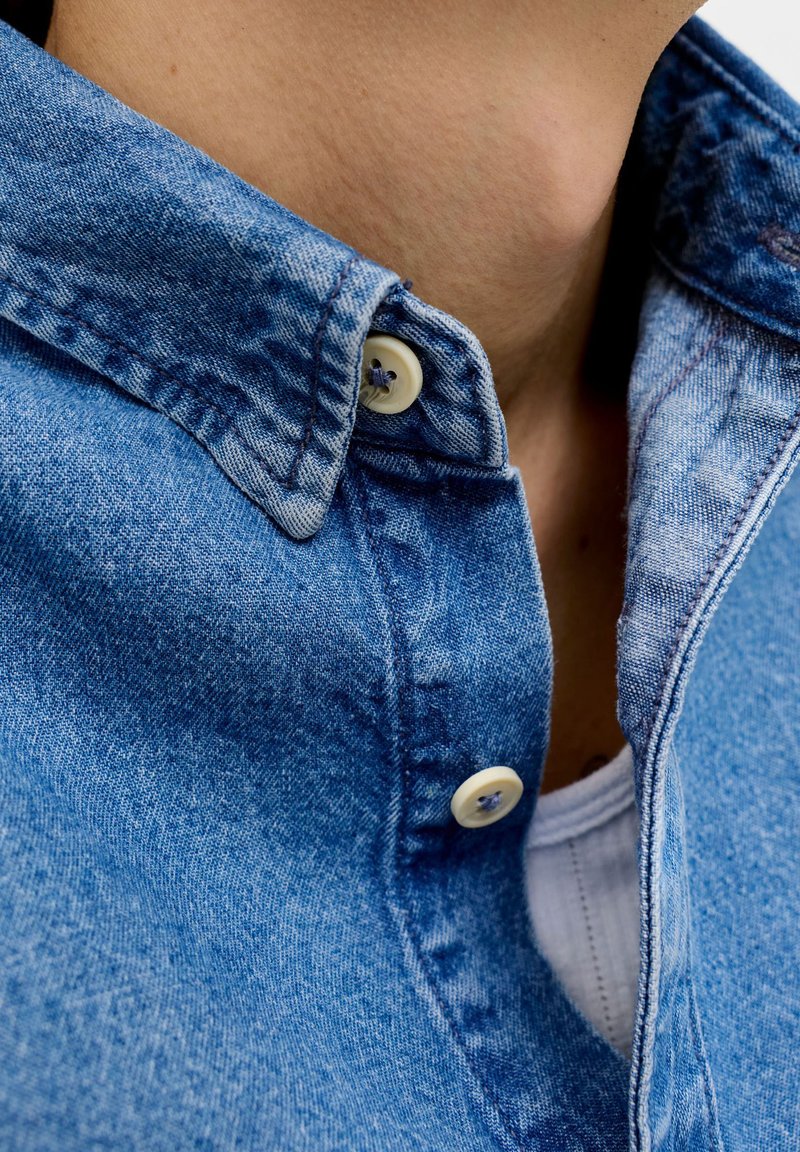 Približni posnetek osebe, ki nosi modro denim srajco z gumbi čez belo spodnjo majico, prikazan je ovratnik in zgornji del prsnega koša.