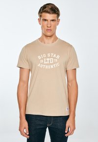 Beežist puuvillane T-särk ümar kaelusega, valge trükitekstiga "BIG STAR 1979 AUTHENTIC", lühikeste varrukatega, kombineeritud tume teksapükstega.