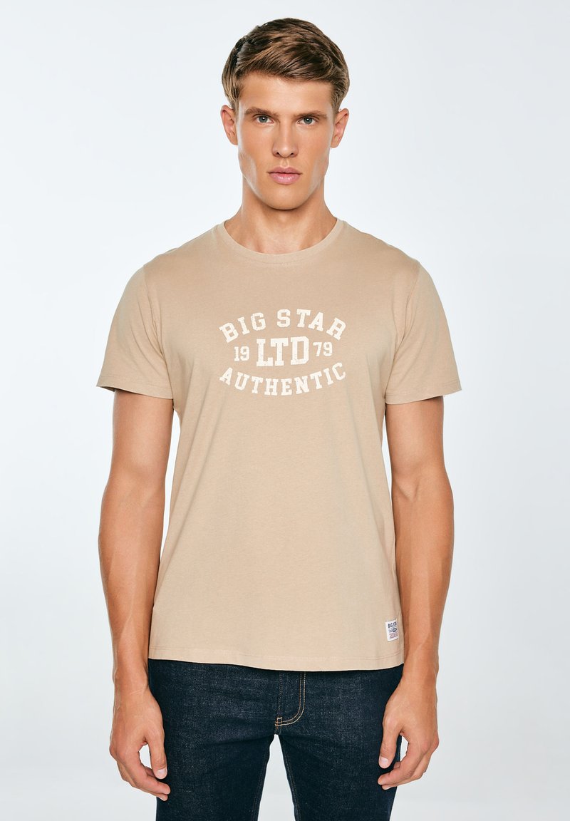 Beežist puuvillane T-särk ümar kaelusega, valge trükitekstiga "BIG STAR 1979 AUTHENTIC", lühikeste varrukatega, kombineeritud tume teksapükstega.