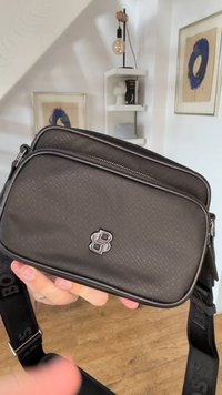 Μαύρη τσάντα crossbody με υφή, διπλό λογότυπο B, ρυθμιζόμενο λουρί, κρατημένη στο χέρι μέσα σε δωμάτιο με κορνιζαρισμένη μπλε τέχνη και λευκά ράφια στο φόντο.