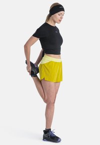 Top deportivo negro, pantalones cortos amarillos con cinturilla elástica y zapatillas negras con acentos morados. El sujeto estira una pierna, sosteniendo un pie.