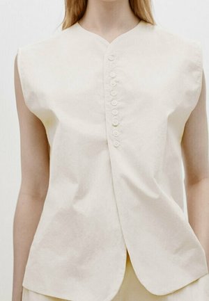 Hihaton beige toppi, jossa epäsymmetrinen etukiinnitys ja pystysuora rivi, jossa yhdeksän pientä nappia, yllä vaaleaihoinen henkilö.