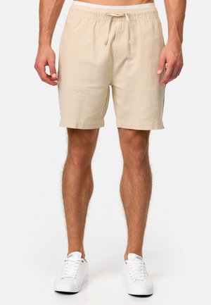 Beige katoenen shorts met een elastische tailleband en trekkoord, met een comfortabele pasvorm en omgeslagen zomen. Draagt wit met sneakers.