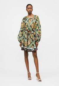 Woman wearing a floral patterned dress with long puffed sleeves, cinched waist by a brown belt, and white high-heeled sandals.