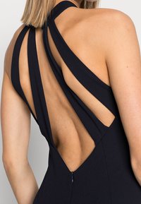 Svart ärmlös klänning med hög halsringning, som har flera urklippsremmar och en öppen rygg, avslutad med dragkedja.