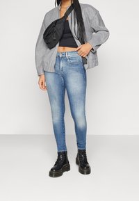 Chaqueta bomber gris, top negro corto, jeans ajustados de mezclilla azul claro y botas negras con cordones. Presenta un bolso negro de bandolera usado en cruz.