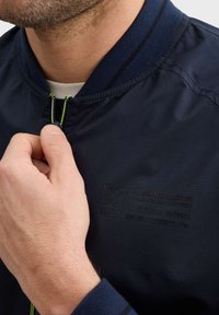 Námořnická bombardér bunda se zipem vpředu, žebrovaným límcem a strukturovanou tkaninou. Obsahuje embosované logo a kontrastní švy.