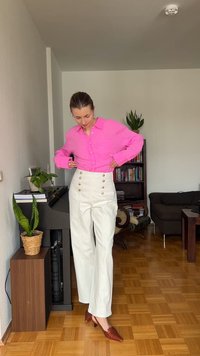 Γυναίκα προσαρμόζει λευκό παντελόνι με ψηλή μέση και χρυσά κουμπιά, φοράει φωτεινό ροζ πουκάμισο και καφετιά παπούτσια με τακούνι σε σαλόνι.