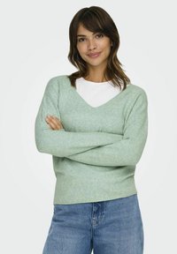 Svetlo zelena v-ratna pulover z mehko teksturo, dolgimi rokavi in sproščenim fitom, v kombinaciji z modrimi kavbojkami.