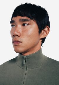 Top verde con texture, con zip e colletto alto. Presenta un dettaglio della zip a contrasto, design aderente e tessuto a coste.