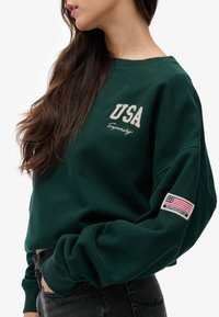 Sudadera verde con "USA" en color crema, con un parche de bandera blanca y roja en la manga. Hecha de material suave y con un corte relajado.