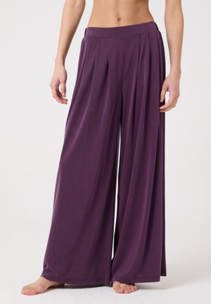 Personne pieds nus portant un pantalon violet taille haute, jambes larges et plissé, se tenant devant un fond blanc uni.
