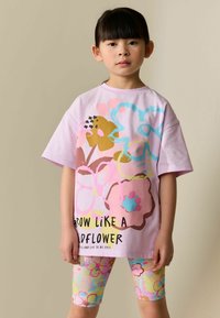 Rozā kokvilnas T-krekls ar ziedu rakstu dzeltenā, zilā un rozā krāsā un uzrakstu "AUGT KĀ SAVVAĻAS ZIEDS". Krekls kombinēts ar krāsainām ziedu legingām.