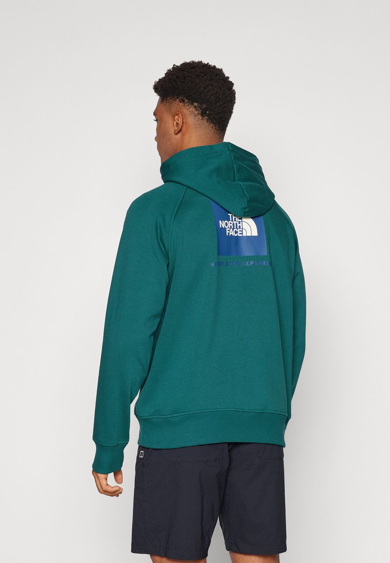 Felpa con cappuccio verde con una toppa con logo sul retro, caratterizzata da una tasca a marsupio e polsini a coste. Indossata con pantaloni corti scuri.
