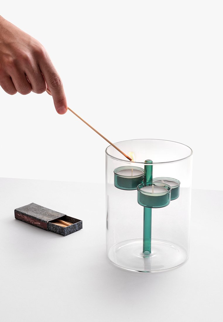 Przezroczysty szklany świecznik z trzema miejscami na tealighty i zielonym wsparciem. Drewniany patyk zapalający świeczkę; zapałki w pobliżu.