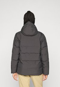 Persoon met gevlochten haar, gekleed in een zwarte gewatteerde jas met capuchon en beige broek, met de rug naar de camera gekeerd tegen een effen grijze achtergrond.