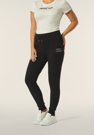 Pantaloni sportivi neri con polsini elasticizzati e tasche, caratterizzati da testo ricamato oro "FREDDY TRAINING." Abbinati a una maglietta bianca e a sneakers bianche.