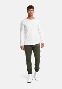 Jeune homme debout, portant une chemise blanche à manches longues, un pantalon cargo vert olive, et des baskets montantes blanches, sur un fond uni.
