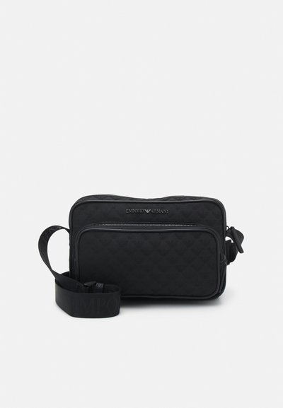 Black quilted fabric crossbody bag with a zip closure, adjustable strap, and front pocket. Features subtle brand logo in silver.