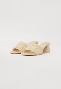 Beige woven slide sandals with an open toe, featuring a chunky heel and a smooth texture. Simple design with a classic look.