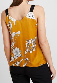 Top senza maniche in satin giallo con spalline nere, caratterizzato da motivi floreali bianchi. L'orlo è leggermente curvato, creando un fit rilassato.