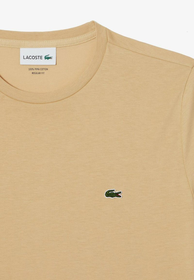 Beżowa koszulka T-shirt z bawełny Pima w regularnym kroju, z małym zielonym logo krokodyla Lacoste wyhaftowanym na lewej piersi.