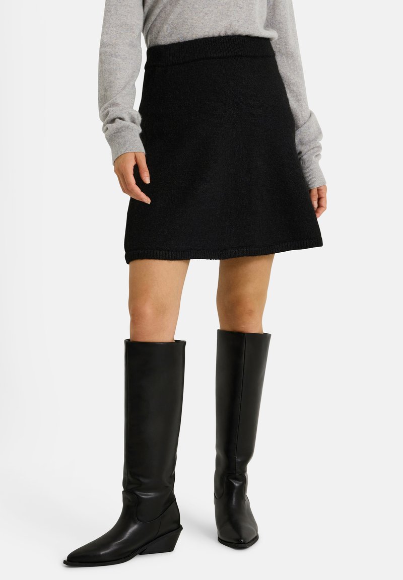 Jupe tricotée noire évasée avec une ceinture ; associée à des bottes en cuir noir jusqu'aux genoux avec un pointu et un talon compensé.