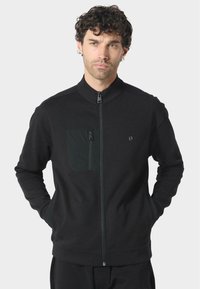 Černá zipová bunda vyrobená z měkké tkaniny, s kapsou na zip na hrudi, vysokým límcem a žebrovanými manžetami. Jednoduchý design, bez vzorů.